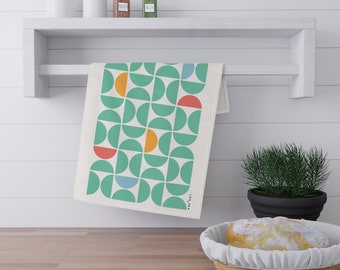 May include: A white kitchen towel with a geometric pattern of green, orange, red, and blue semi-circles. The towel is hanging on a white shelf with jars of spices on the shelf. The text "Leila" is printed on the towel.