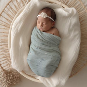 May include: A newborn baby is sleeping peacefully in a woven basket, swaddled in a light blue muslin wrap with a white and gray cloud pattern. The baby is wearing a white headband with a small flower.