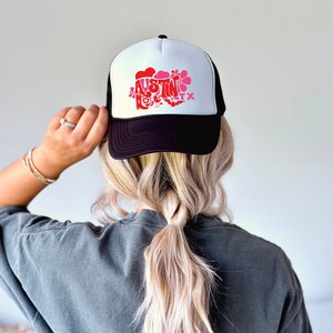 Può includere: Cappellino da camionista nero e bianco con una grafica rosa e rossa che dice "Austin TX" con un fiore, un uccello e un pipistrello.
