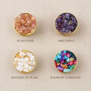 May include: Four circular gold-toned metal trays filled with small tumbled stones. The top left tray contains orange sunstone, the top right contains purple amethyst, the bottom left contains white mother of pearl, and the bottom right contains multicolored rainbow gemstones.