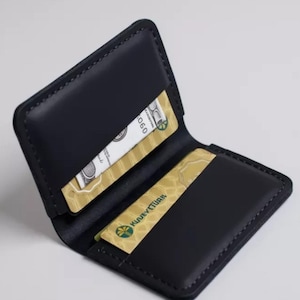 May include: A black leather bifold wallet with visible stitching along the edges. The wallet is open, revealing two interior slots. One slot holds a white paper bill with visible text, and the other holds a gold-coloured card with the text KUVEYTTÜRK.