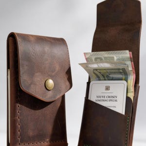 Pode incluir: Dois porta-cartões de couro castanho com costuras visíveis. Um está fechado com um botão de pressão de latão, enquanto o outro está aberto, revelando notas e um cartão branco com o texto "YOU'VE CHOSEN SOMETHING SPECIAL" impresso.