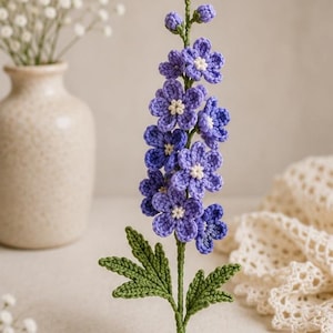 May include: A handcrafted crochet delphinium flower stem featuring multiple vibrant purple blossoms with small white centers, green textured leaves, and a sturdy green stem, topped with several small, unopened purple flower buds.
