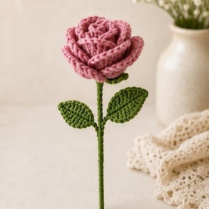 Può includere: Una rosa all'uncinetto realizzata a mano con un fiore rosa polvere dai petali stratificati, un lungo stelo verde e due foglie verdi attaccate allo stelo. Il fiore è realizzato in filato con una trama lavorata a maglia visibile ovunque.