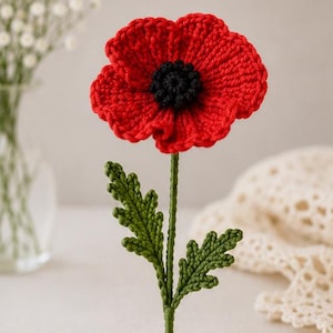 May include: A handcrafted crochet poppy flower featuring vibrant red petals and a textured black center. The flower is attached to a long, thin green stem with two detailed, serrated green leaves extending from the sides of the stalk.