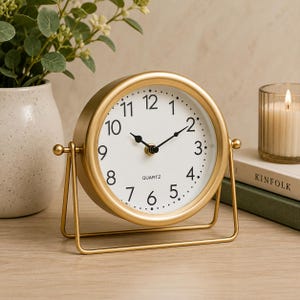 May include: A round table clock with a gold-toned metal frame and a white face. The clock features black Arabic numerals from 1 to 12, black hour and minute hands, and the word QUARTZ printed in black. It is mounted on a gold-toned metal stand.