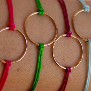 May include: Seven bracelets with gold circle charms on a wrist. The bracelets are made with different colored cords: pink, green, burgundy, light blue, hot pink, and dark green.