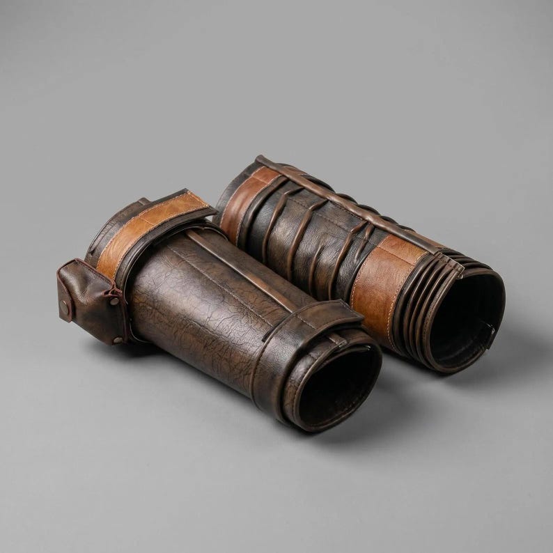 May include: A pair of dark brown and tan forearm guards featuring a textured surface, horizontal strap detailing, and a small attached pouch on one side. These cylindrical arm protectors are constructed with layered panels and secure closures.