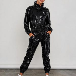 Könnte beinhalten: Ein schwarzer Trainingsanzug aus glänzendem Vinyl, bestehend aus einer langärmeligen Reißverschlussjacke mit Stehkragen und einer passenden Hose mit elastischen Bündchen an Taille und Knöcheln. Das Material hat eine glänzende Oberfläche und wird mit weißen Sneakern kombiniert.