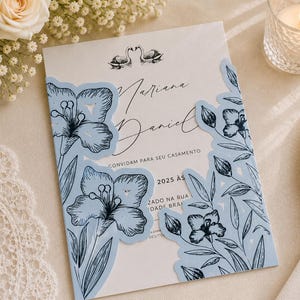 Pode incluir: Um convite de casamento branco com o texto preto Mariana e Daniel e uma ilustração de cisnes. O cartão está inserido numa capa de papel azul-claro cortada a laser, decorada com desenhos florais a traço preto e motivos botânicos.