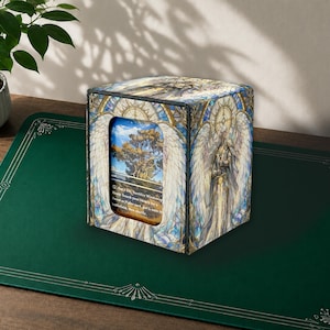 May include: A square deck box featuring a stained glass art style with an angel holding a sword. The box has a central cutout window displaying a Windswept Heath card with text that reads: Pay 1 life, Sacrifice Windswept Heath: Search your library for a Forest or Plains card, put it onto the battlefield, then shuffle.