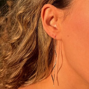 May include: A long, slender silver-tone threader earring featuring a delicate, wavy metallic wire design that hangs vertically from the earlobe, creating a minimalist and fluid appearance with a polished, reflective surface finish.