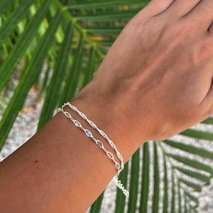 May include: A double-strand silver bracelet featuring one chain with a twisted, rope-like texture and a second chain adorned with small, light blue, oval-shaped stones set in silver metal links, worn around the wrist.