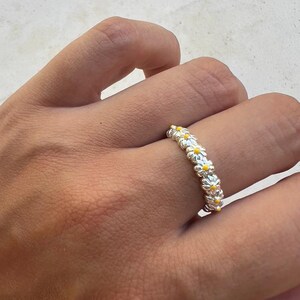 Puede incluir: Un anillo de plata que presenta una banda continua de pequeñas flores de margarita esculpidas. Cada flor individual tiene pétalos de plata metálica y un pequeño centro de color amarillo brillante, creando un patrón floral repetido alrededor del anillo.