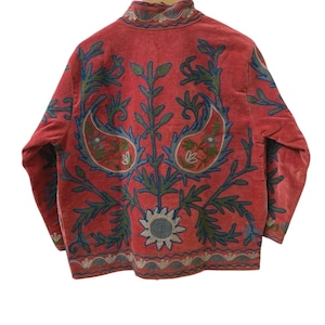 May include: A long-sleeved coral-colored jacket featuring intricate embroidery in shades of blue, green, and cream. The design includes large paisley motifs, floral patterns, and a sun-like emblem at the bottom, all displayed on a textured fabric.