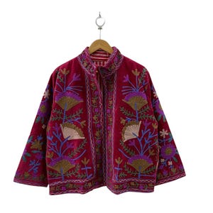 Pode incluir: Um casaco magenta com intrincados bordados florais multicoloridos em tons de roxo, azul e dourado. A peça tem gola alta, mangas compridas e uma abertura frontal, exibindo padrões de costura detalhados em toda a superfície.