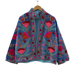 Pode incluir: Uma jaqueta azul com bordados florais vibrantes em tons de rosa, laranja e vermelho. A peça possui gola alta, mangas compridas e um design aberto na frente. O forro interno é visível e exibe um padrão de listras azul claro e branco.