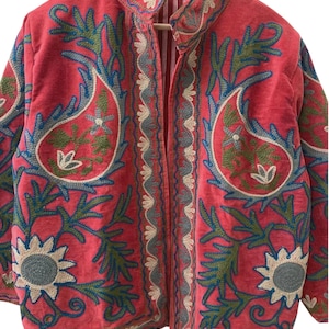 May include: A vibrant pink jacket featuring intricate embroidery in shades of blue, green, and cream. The design showcases large paisley motifs, floral patterns, and decorative borders along the front opening and hem, with a striped inner lining.