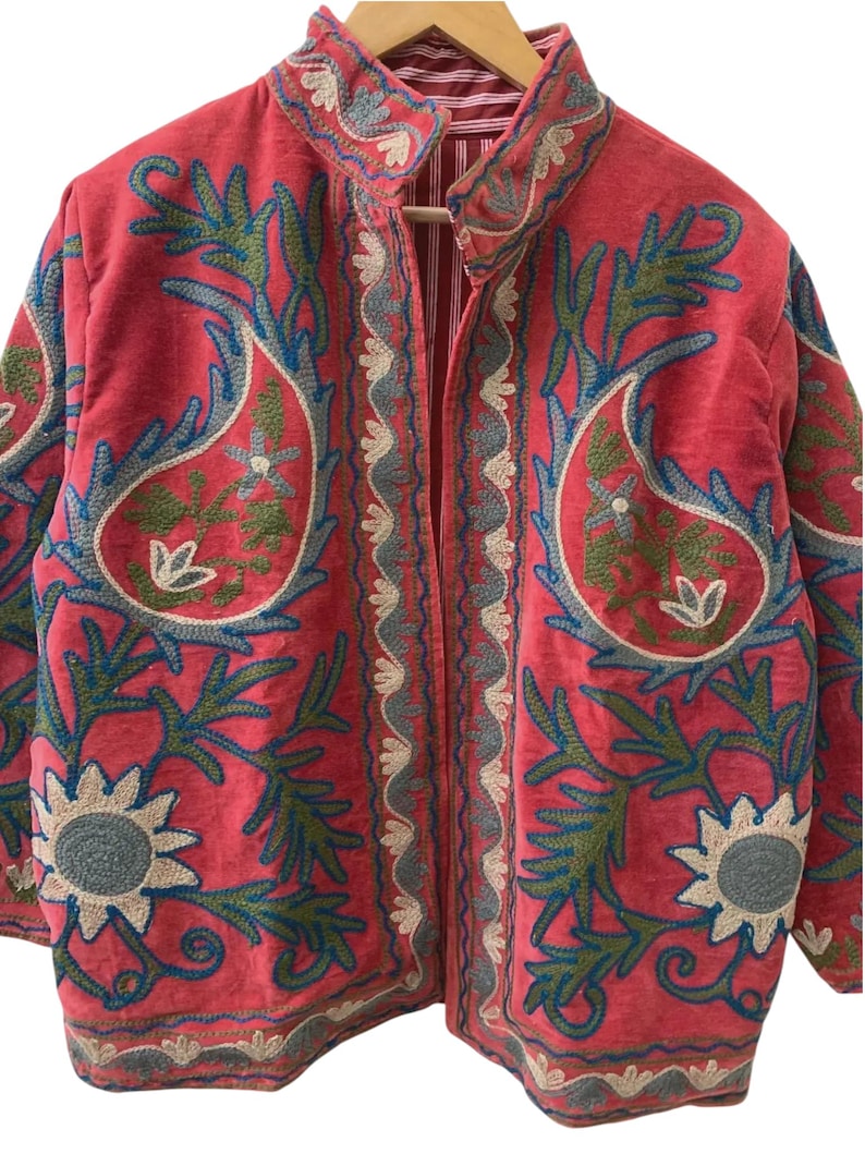 May include: A vibrant pink jacket featuring intricate embroidery in shades of blue, green, and cream. The design showcases large paisley motifs, floral patterns, and decorative borders along the front opening and hem, with a striped inner lining.
