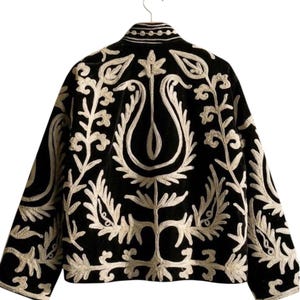 Puede incluir: Una chaqueta negra con intrincados bordados blancos en un patrón simétrico de flores y hojas en la espalda y las mangas. La prenda tiene un cuello alto con líneas blancas horizontales y mangas largas, presentada sobre una percha.