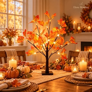 Puede incluir: Un árbol decorativo de mesa con ramas oscuras adornadas con hojas de arce naranjas, rojas y amarillas, iluminadas por pequeñas luces LED de color blanco cálido, sobre una base cuadrada, con el texto Elora Wooden en la esquina superior.