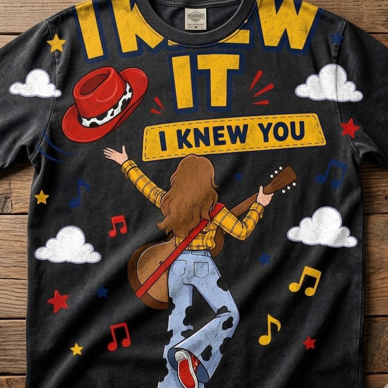 May include: A black t-shirt featuring a graphic of a person with long brown hair wearing a yellow plaid shirt and cow-print bell-bottom trousers, playing an acoustic guitar. The text reads I KNEW IT and I KNEW YOU, with a red cowboy hat, clouds, and stars.
