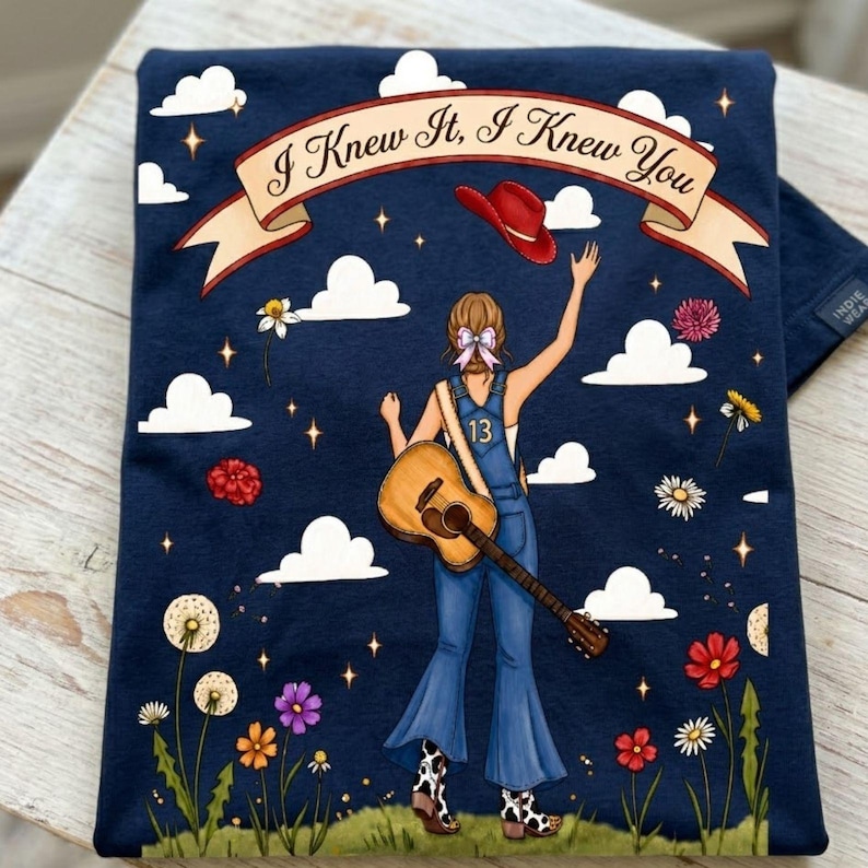 May include: A navy blue t-shirt featuring a graphic of a person in blue overalls with the number 13, holding an acoustic guitar and a red cowboy hat. The design includes white clouds, colorful flowers, and a banner with the text I Knew It, I Knew You.