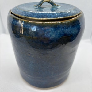May include: A cylindrical ceramic jar with a glossy, deep blue glaze and a matching lid. The lid features a small, arched handle at the centre. The surface of the jar shows subtle variations in blue tones, ranging from dark navy to lighter cobalt.