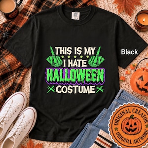 May include: A black short-sleeve t-shirt featuring a graphic with green skeleton hands making middle finger gestures. The text reads THIS IS MY I HATE HALLOWEEN COSTUME in white and green lettering with a purple outline on the shirt.