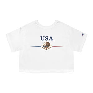 Pode incluir: Um top curto branco de manga curta com o texto USA em letras azuis acima de um emblema de águia-de-cabeça-branca. Uma linha azul fina estende-se para a esquerda e uma linha vermelha fina para a direita do emblema. Um pequeno logótipo Champion na manga.