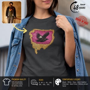 Peut inclure: Un t-shirt gris anthracite avec un motif central représentant un oiseau noir en vol, sur un fond magenta texturé bordé d'une bordure dorée vieillie. Le t-shirt est porté sous une veste en jean bleue avec des boutons métalliques visibles.
