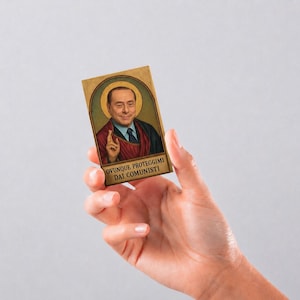 May include: A rectangular card featuring a portrait of a man in a suit with a golden halo behind his head, styled like a religious icon. The man is making a hand gesture, and the text at the bottom reads, "OVUNQUE PROTEGGIMI DAI COMUNISTI."