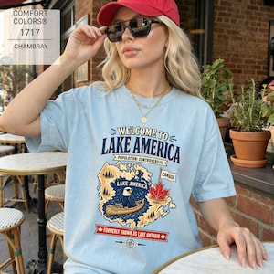 May include: A light blue t-shirt featuring a graphic of a map labeled Lake America, a bald eagle in a canoe, a red maple leaf, and the text Welcome to Lake America, Population: Controversial, Canada, and Formerly Known as Lake Ontario.
