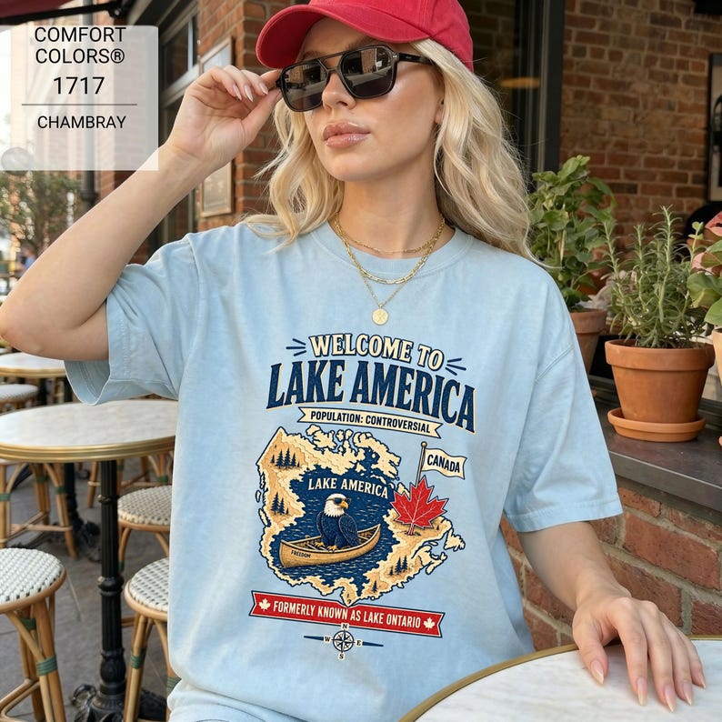May include: A light blue t-shirt featuring a graphic of a map labeled Lake America, a bald eagle in a canoe, a red maple leaf, and the text Welcome to Lake America, Population: Controversial, Canada, and Formerly Known as Lake Ontario.