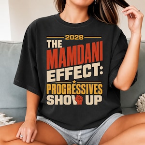 以下が含まれることがあります： 黒の半袖Tシャツ。オレンジ、黄色、白の文字で「2028 THE MAMDANI EFFECT: PROGRESSIVES SHOW UP」とプリントされており、小さなオレンジ色の星と、SHOWの「O」の代わりに拳を突き上げたアイコンがデザインされています。