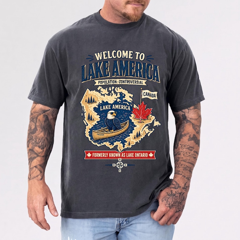 May include: A charcoal gray short-sleeve t-shirt featuring a graphic print that reads "WELCOME TO LAKE AMERICA POPULATION: CONTROVERSIAL". The design includes a map, a bald eagle in a canoe, a red maple leaf, and "FORMERLY KNOWN AS LAKE ONTARIO".