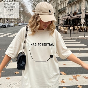 Op de afbeelding: Een ivoorkleurig t-shirt met de zwarte tekst I HAD POTENTIAL boven een afbeelding van een gebogen lijn en een kleine zwarte cirkel. Het shirt wordt gecombineerd met een beige honkbalpet met een klein zwart logo en een zwarte schoudertas met ketting.