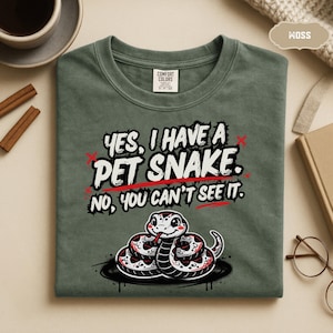 May include: A moss green crew neck t-shirt featuring a graphic of a coiled snake and the text, Yes, I have a pet snake. No, you can't see it. The design uses white, black, and red ink. The shirt has a Comfort Colors brand label at the neck.