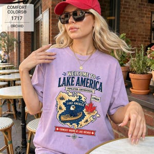 May include: A light purple t-shirt featuring a graphic print with the text Welcome to Lake America, Population: Controversial, Canada, and Formerly Known as Lake Ontario. The design includes a bald eagle in a canoe, a maple leaf, and a map.