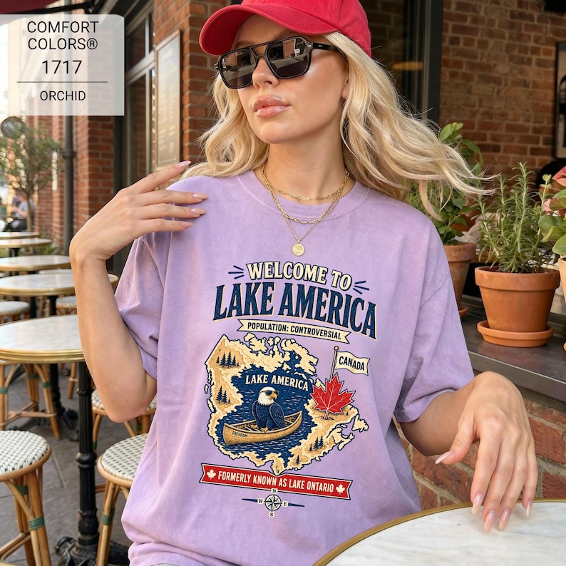May include: A light purple t-shirt featuring a graphic print with the text Welcome to Lake America, Population: Controversial, Canada, and Formerly Known as Lake Ontario. The design includes a bald eagle in a canoe, a maple leaf, and a map.