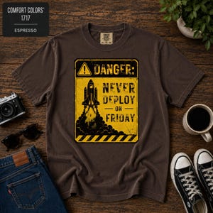 May include: A dark brown short-sleeve t-shirt featuring a yellow rectangular graphic with a black border. The graphic displays a rocket ship, a warning triangle, and the text DANGER: NEVER DEPLOY ON FRIDAY with black hazard stripes.