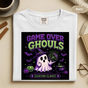 May include: A white t-shirt featuring a pixel art graphic with the text "GAME OVER GHOULS" and "[CUSTOM CLASS]". The design shows a ghost wearing a purple witch hat, holding a twenty-sided die, surrounded by bats, pumpkins, and a cauldron.