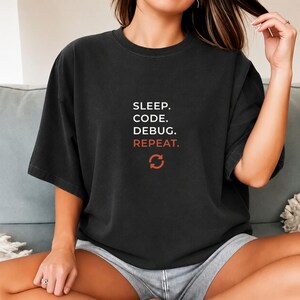 Op de afbeelding: Een zwart t-shirt met korte mouwen voorzien van de witte tekst SLEEP. CODE. DEBUG. en de oranje tekst REPEAT. eronder. Een oranje cirkelvormig verversingsicoon is onder de tekst geplaatst. Het t-shirt heeft een ronde hals en een ruime pasvorm.