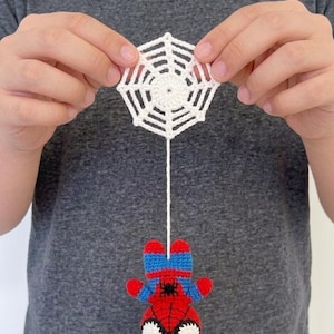 Puede incluir: Un pequeño adorno de ganchillo hecho a mano que presenta un personaje de superhéroe rojo y azul con un emblema de araña negra en el pecho, colgado de una telaraña de ganchillo blanca mediante un hilo fino. El personaje tiene grandes ojos blancos.