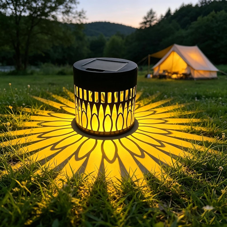 May include: A cylindrical solar-powered lantern with a black frame and a cutout pattern that casts a bright, warm yellow sunburst light projection onto the surrounding grass, featuring a flat solar panel on the top surface of the device.