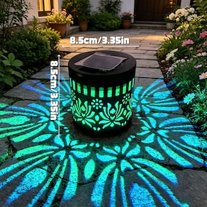 May include: A cylindrical black solar garden light with a cutout floral pattern that projects a bright turquoise light onto the ground. The device features a square solar panel on top and measures 3.35 inches in height and 3.35 inches in diameter.