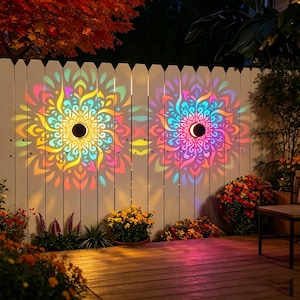 Pode incluir: Duas luminárias de parede circulares para exterior montadas numa cerca de madeira branca, projetando padrões de luz multicoloridos tipo mandala em tons de amarelo, laranja, rosa e azul sobre a superfície durante a noite.