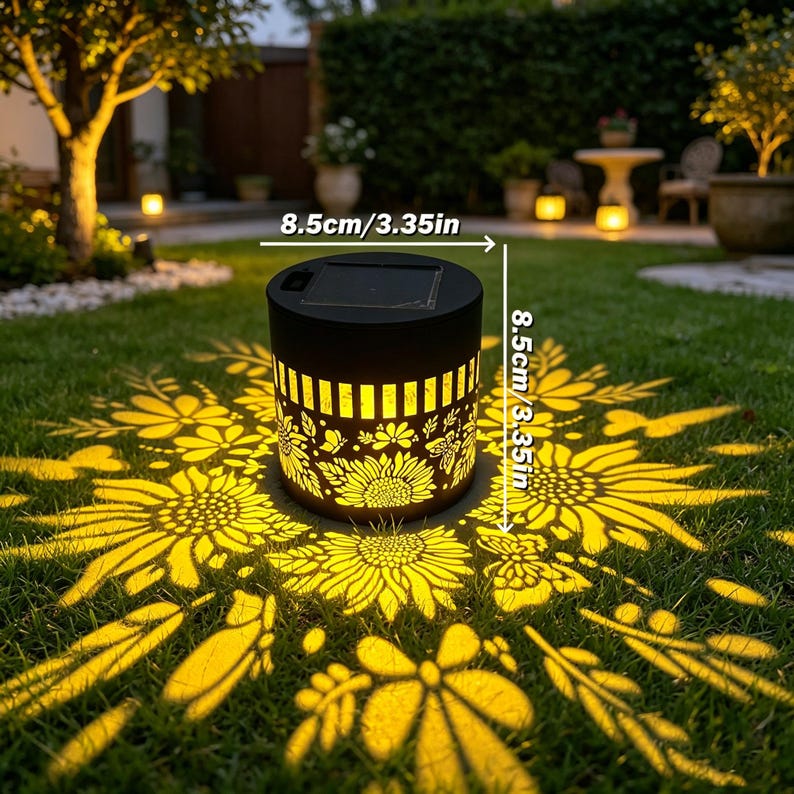 May include: A cylindrical black solar garden light with a cutout pattern of sunflowers and butterflies. The light projects a bright yellow floral design onto the grass. It measures 3.35 inches in height and 3.35 inches in diameter.
