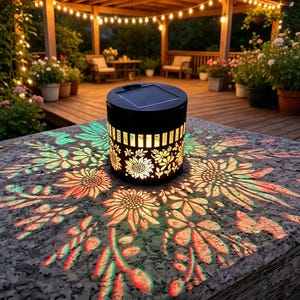 May include: A cylindrical black solar lantern with a cut-out floral pattern that projects a glowing sunflower and leaf design onto the surface below. The top of the lantern features a square solar panel, and the sides emit a warm light.