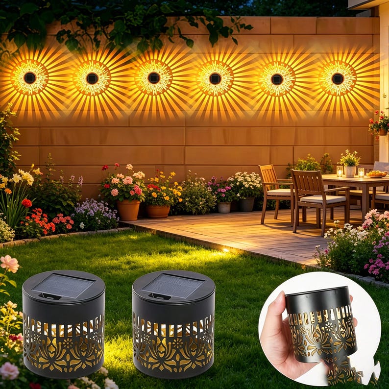 May include: Two cylindrical black solar-powered outdoor lights featuring intricate laser-cut floral patterns on the sides and a square solar panel on top, shown both placed on a lawn and held in a hand to display the detailed metal cutout design.